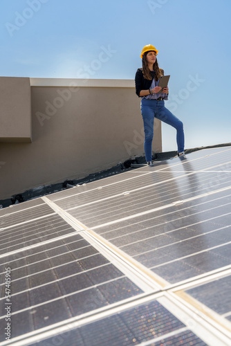 Ingegnere donna con caschetto protettivo giallo verifica le condizioni di un impianto fotovoltaico sul tetto di una casa con un tablet in mano