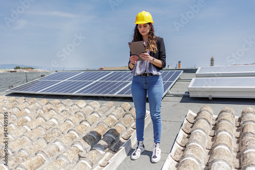 Ingegnere donna con caschetto protettivo giallo verifica le condizioni di un impianto fotovoltaico sul tetto di una casa con un tablet in mano photo
