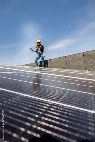 Ingegnere donna con caschetto protettivo giallo verifica le condizioni di un impianto fotovoltaico sul tetto di una casa con un tablet in mano