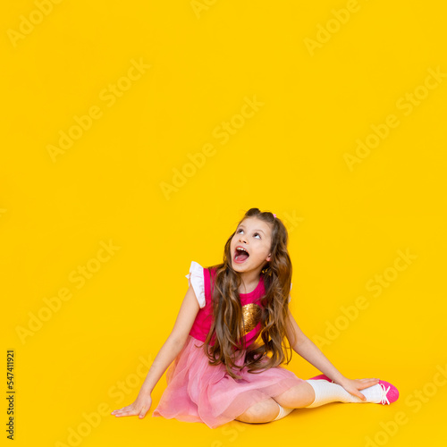 A little girl is sitting and looking up at your advertisement on a yellow isolated background. A child with long wavy hair in a pink dress is smiling broadly.