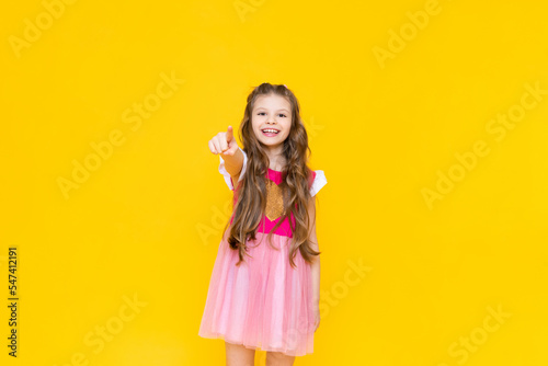 A little princess in a pink dress with curly hair smiles broadly and points forward with her index finger. A beautiful, charming child on a yellow isolated background.