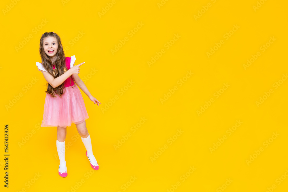 A little girl points to your ad on a yellow isolated background. A beautiful child in a pink dress points to an empty place and smiles broadly. A little girl points to your ad on a yellow isolated background. A beautiful child in a pink dress points to an empty place and smiles broadly.