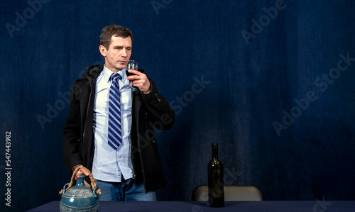 White man in strict business suit stands against dark background. He holds glass of wine in his hand and prepares to drink it. Start of drinking.