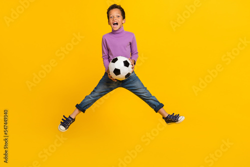 Full size photo of overjoyed energetic boy jumping hands hold football isolated on yellow color background © deagreez