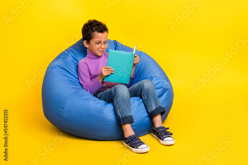 Full size portrait of smart schoolboy sitting comfy bag read book isolated on yellow color background