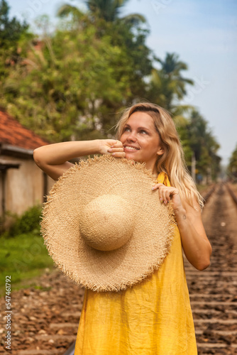 Young woman wearing yellow dress with straw hat standing on railway track at rural, looking away. Lady model in summer wear in tropical countryside backdrop. Travel vacation concept. Copy text space