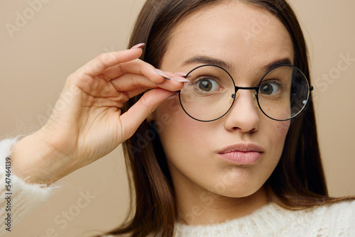 an attractive woman is standing on a light background in a white sweater holding her glasses with the fingers of one hand and slightly pouting her lips looking forward with wide eyes