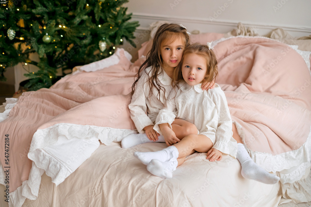 Two little curly-haired sisters are sitting on the bed in their nightgowns on the morning after Christmas. Soft pink bedding on the bed