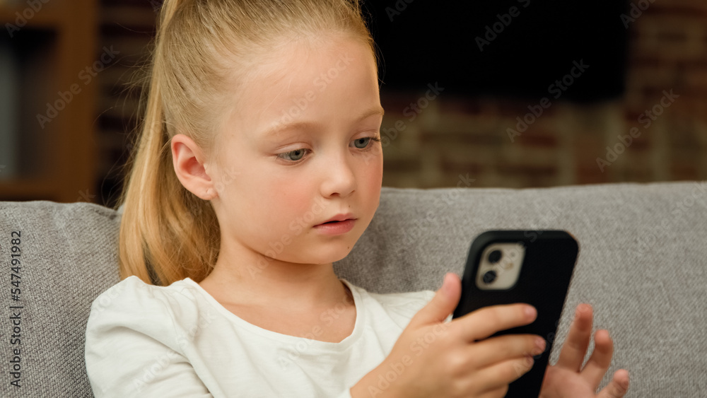 Adorable little kid girl cute child sitting on cozy couch alone in living room looking at gadget screen using smartphone software application playing mobile game watching video on phone relax leisure Adorable little kid girl cute child sitting on cozy couch alone in living room looking at gadget screen using smartphone software application playing mobile game watching video on phone relax leisure