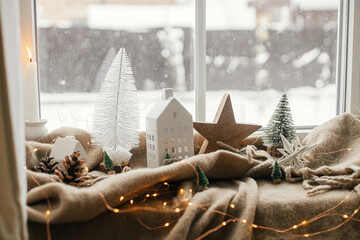 Christmas still life, cozy winter home. Stylish christmas decorations, tree, lights, little house and wooden star on cozy blanket on windowsill. Merry Christmas! Atmospheric scandinavian mood