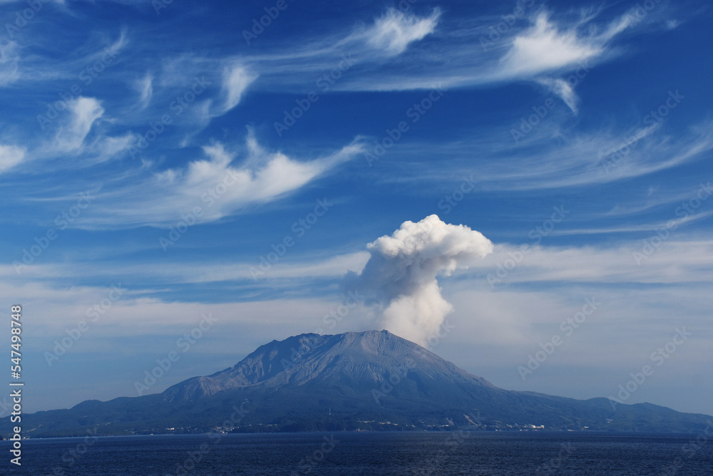 Sakurajima, an active volcano in Japan, rises above the ocean under a bright blue sky. White smoke emerges from its peak, surrounded by wispy clouds, creating a dramatic landscape. Sakurajima, an active volcano in Japan, rises above the ocean under a bright blue sky. White smoke emerges from its peak, surrounded by wispy clouds, creating a dramatic landscape.