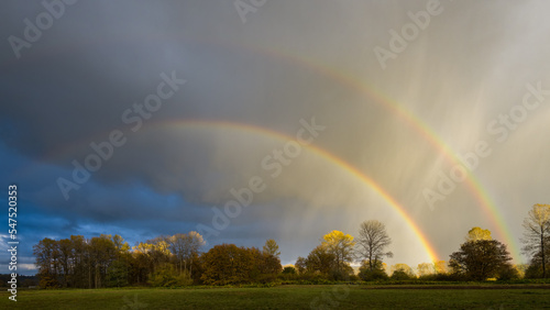 Double rainbow passing through rain shafts above rural countryside in fall