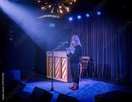 Poet Deborah-Anne Tunney reading from A Different Wolf on a nightclub stage, stage lights, spot lights, piano, microphone and microphone stand
