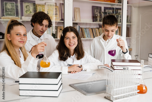Schoolchildren studying at chemistry lesson in classrom. Pupils writing in notebook  holding flasks with liquid for experiments and have fun together. School education.