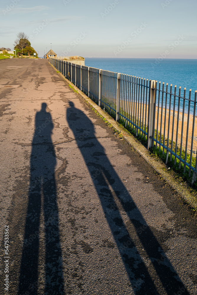 The silhouette of  two people walking along a promenade next to the sea.