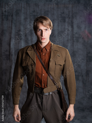 A young guy in military-style clothes, a brown flight jacket and breeches with suspenders. Posing in the studio on a gray background, close-up