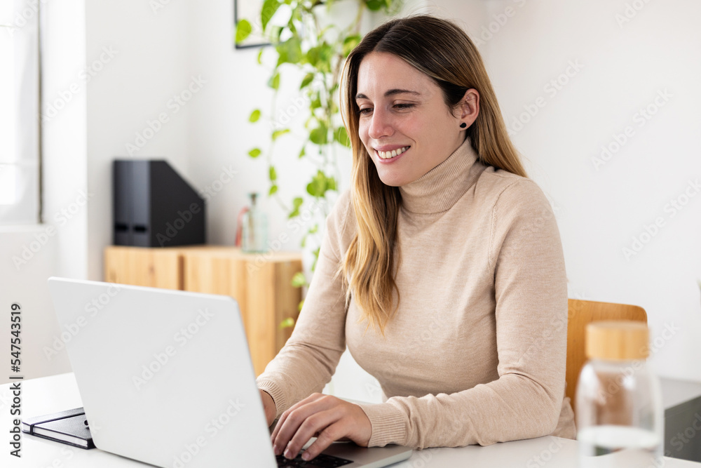 Happy young smiling woman working on laptop from home office. Business female entrepreneur sitting on desk typing on computer Happy young smiling woman working on laptop from home office. Business female entrepreneur sitting on desk typing on computer