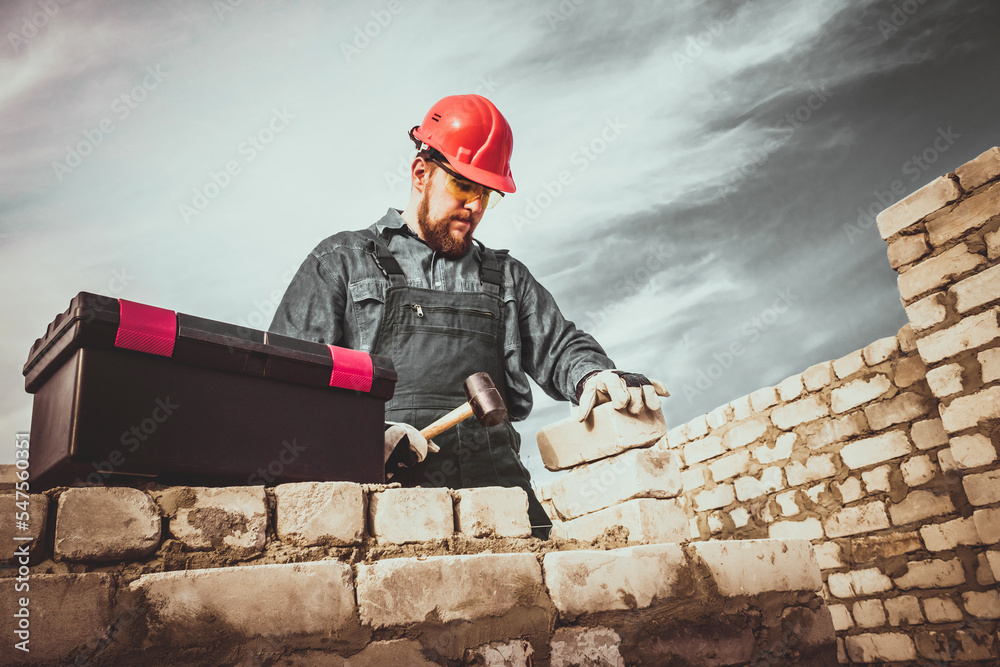 Photo of male construction site worker in hardhat and overalls uniform building wall with toolbox and hammer on blue skies background.