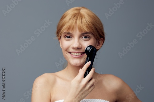 a happy, joyful woman stands on a gray background wrapped in a towel and does a facial massage with a black electric massager