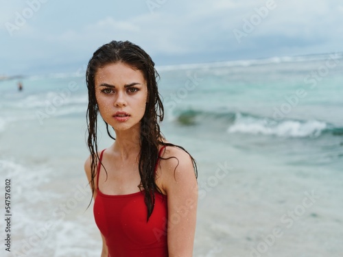 Beautiful woman on the beach by the sea in a red swimsuit looks at the camera