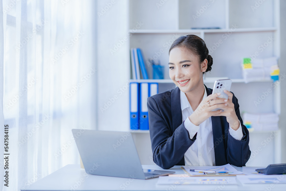 Asian businesswoman using smartphone and laptop to contact customers, calculate with calculator on desk, reading financial graphs, planning, analyzing, calculating market data in corporate office.