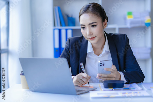 Asian businesswoman using smartphone and laptop to contact customers, calculate with calculator on desk, reading financial graphs, planning, analyzing, calculating market data in corporate office. © ArLawKa