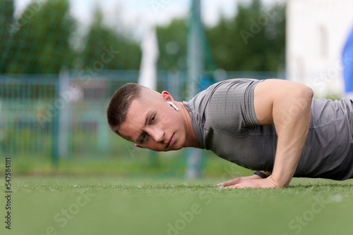 Young athlete does push-ups in an outdoor stadium, listening to music with headphones. Muscular athlete is engaged in sports, performs push-ups on a modern artificial green surface of the stadium