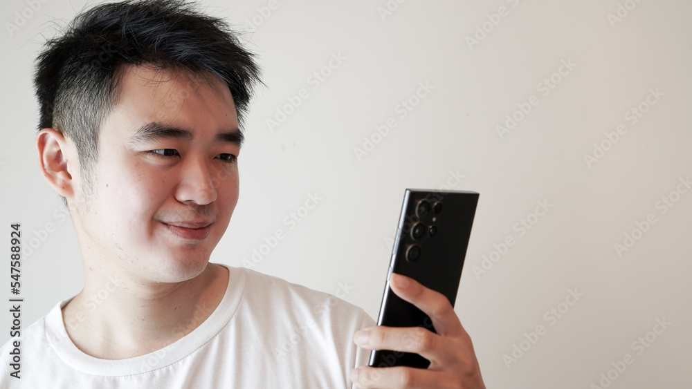 an asian man wearing plain shirt was holding his smartphone or cellphone, reading and thinking of an action while touching his chin isolated over a white background an asian man wearing plain shirt was holding his smartphone or cellphone, reading and thinking of an action while touching his chin isolated over a white background