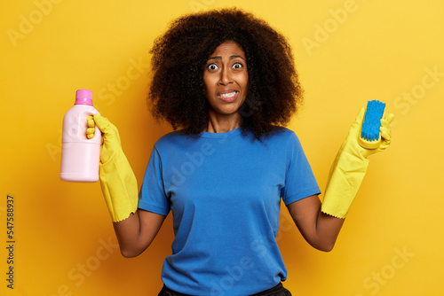 Obraz na płótnie Unhappy dark skinned housewife, wearing yellow gloves and blue t-shirt, holds a