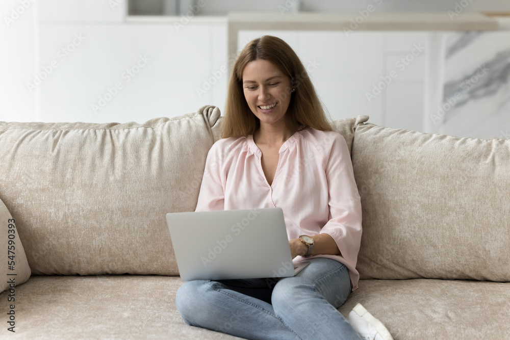 Smiling attractive woman sitting on sofa using laptop, female communicating or working on-line at home, typing on computer, enjoying writing blog or chatting with friends in social media network. Tech Smiling attractive woman sitting on sofa using laptop, female communicating or working on-line at home, typing on computer, enjoying writing blog or chatting with friends in social media network. Tech