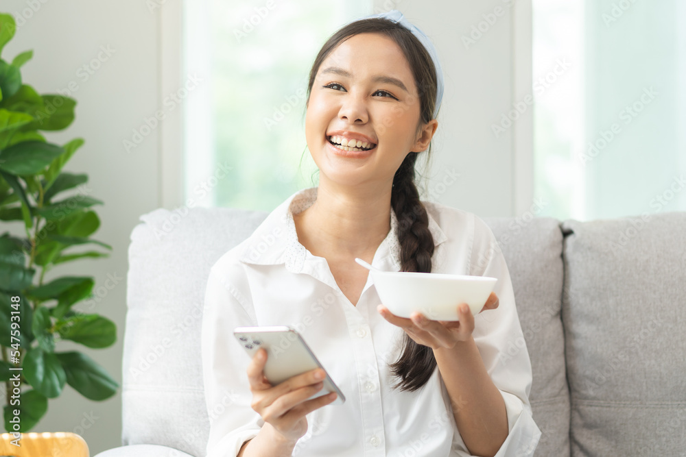 Healthy lifestyle, wellness food asian young woman hand using smartphone sitting relaxed on couch at home, eat tasty yogurt, dairy product meal. Snack tasty on diet nutrition delicious natural yoghurt Healthy lifestyle, wellness food asian young woman hand using smartphone sitting relaxed on couch at home, eat tasty yogurt, dairy product meal. Snack tasty on diet nutrition delicious natural yoghurt