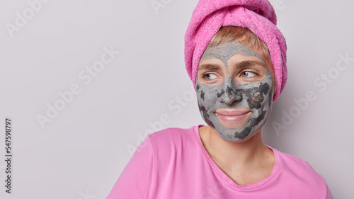 Horizontal shot of thoughtful woman applies beauty clay mask on face focused aside undergoes skin care procedures wears pink t shirt nd bath towel wrapped on head isolated over grey background