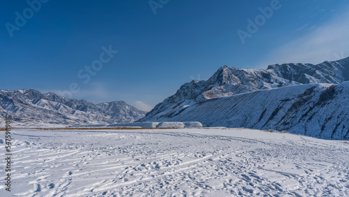 Footprints, car tires, snowmobiles are visible on the snow-covered high-altitude plateau. A picturesque mountain range with sharp peaks against the blue sky. Altai