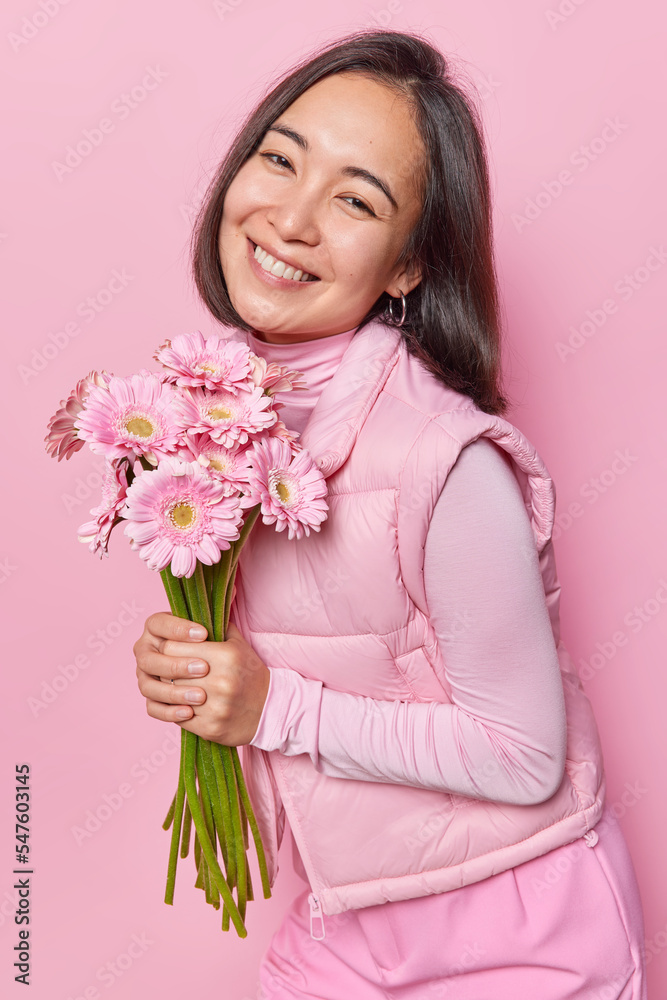 Vertical shot of happy Asian woman receives flowers from husband holds bouquet of gerberas smiles happily dressed in casual jumper and vest isolated over pink background. Special occasion concept