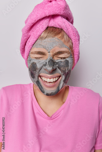 Overjoyed female model laughs joyfully keeps eyes closed smiles toothily applies nourishing clay mask being in good mood wears pink t shirt bath towel wrapped on head. Beauty procedures concept © wayhome.studio 
