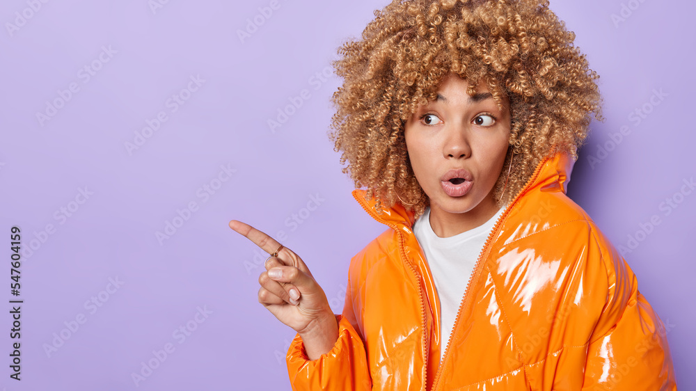 Surprised curly haired woman feels amazed discusses with product dressed in orange jacket shows blank wall for promotional information demonstrate something astonishing isolated on purple background.