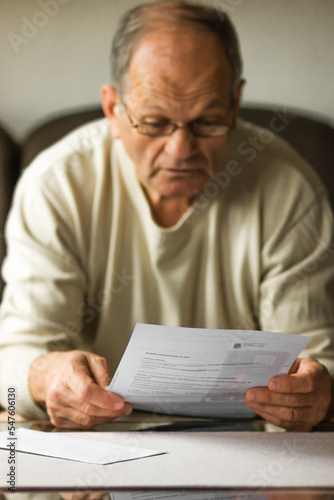 Worried senior man in white sweater and glasses reading utility bill in period of energy crisis. High heating prices.