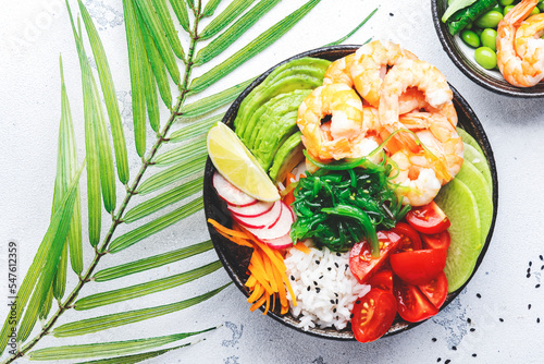 Hawaiian poke bowl with shrimp, avocado, radish, carrot, tomato, chuka seaweed and white rice. Soy sauce, lime and sesame dressing. White table background, top view