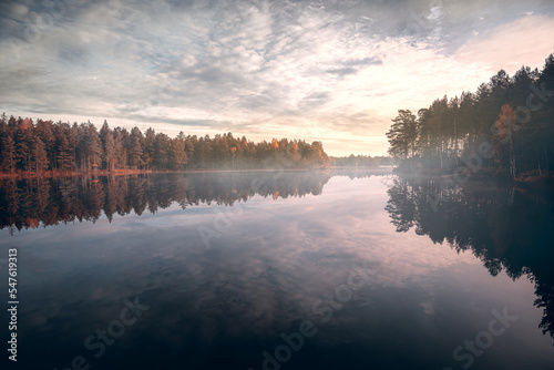 Earlt morning view over forest lake an early fall morning just after the sun rises with light mist over the lake and the trees and sky refelcting in the calm water surface  © imagesbystefan
