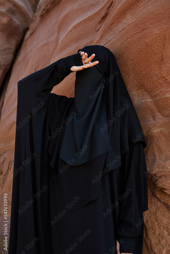Muslim woman in black national clothes and hijab hiding her face with hand in canyon 