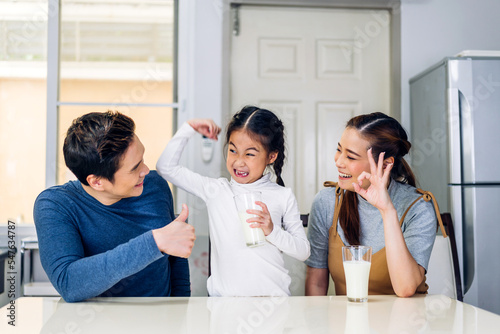 Portrait of enjoy happy love asian family father and mother with little asian girl smiling and having protein breakfast drinking and hold glasses of milk at table in kitchen.