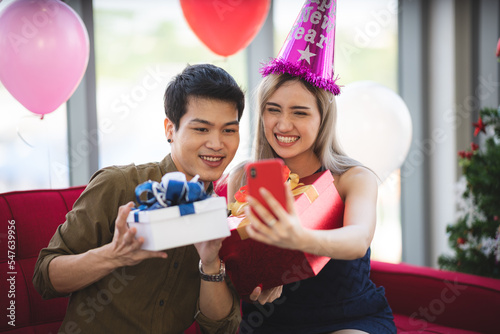 Cheerful young couple celebrating new year by taking selfie on smartphone wearing hats and balloon at home