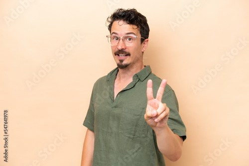 Young caucasian man isolated on beige background smiling and showing victory sign