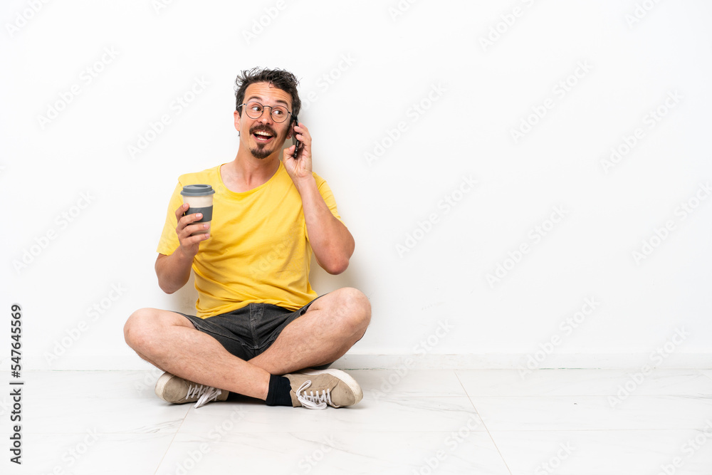 Fototapeta premium Young caucasian man sitting on the floor isolated on white background holding coffee to take away and a mobile