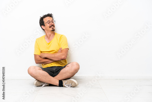 Young caucasian man sitting on the floor isolated on white background with arms crossed and happy