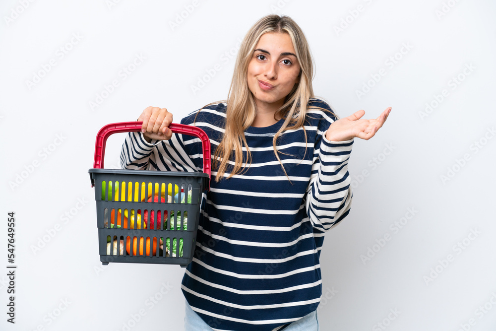 Young caucasian woman holding a shopping basket full of food isolated on white background having doubts while raising hands Young caucasian woman holding a shopping basket full of food isolated on white background having doubts while raising hands