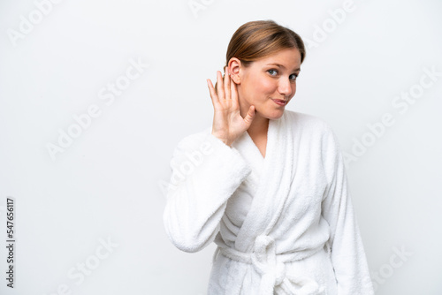 Young pretty blonde woman in bathrobe isolated on white background listening to something by putting hand on the ear