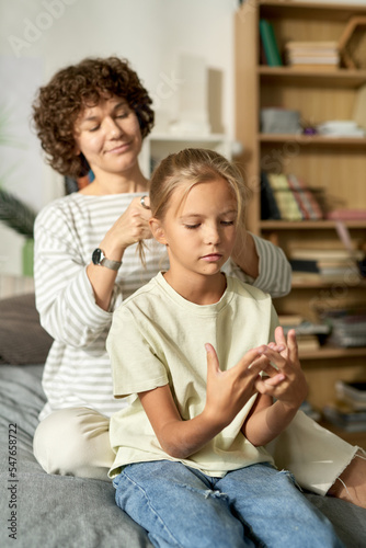Young mother making hairstyle to her daughter while they sitting on bed in bedroom