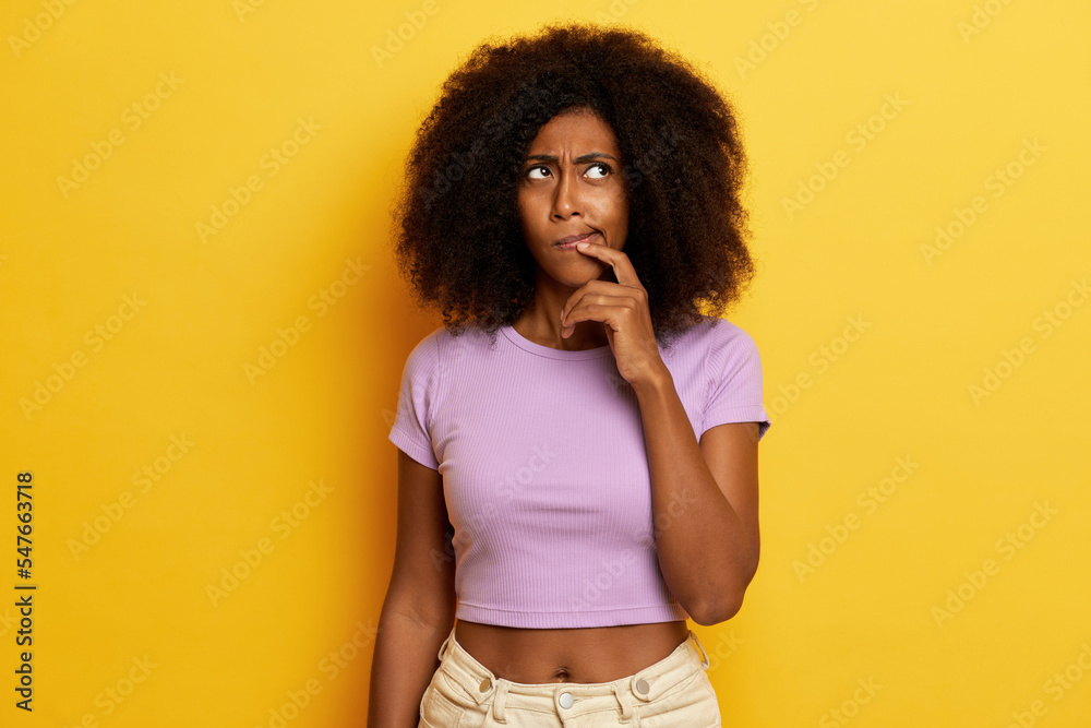 Photo of dreamy dark skinned young woman with afro hair, looks thoughtfully aside, keeps finger near underlip, dressed in purple T-shirt and jeans, posing over yellow wall Photo of dreamy dark skinned young woman with afro hair, looks thoughtfully aside, keeps finger near underlip, dressed in purple T-shirt and jeans, posing over yellow wall