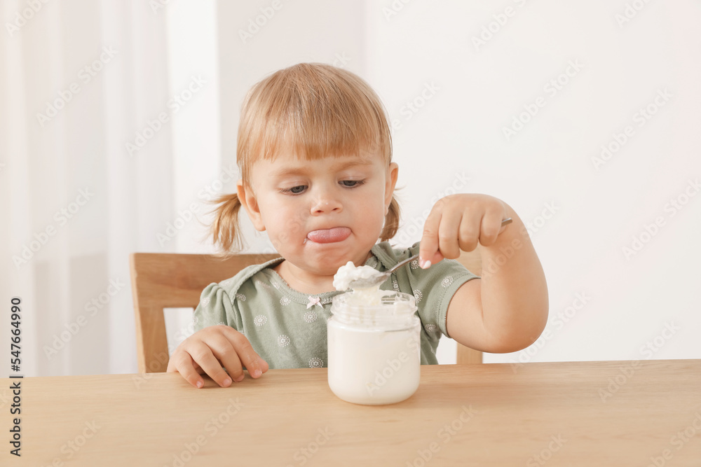 Cute little child eating tasty yogurt with spoon at wooden table indoors Cute little child eating tasty yogurt with spoon at wooden table indoors